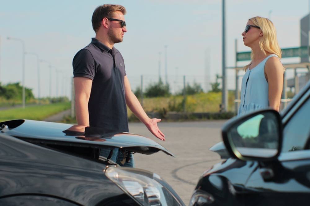 Man and woman drivers shake hands after a small road accident. In the foreground there are two cars.